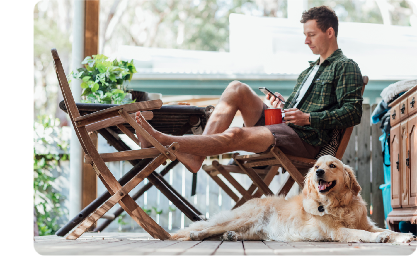 Man relaxing on a chair on his front porch looking at his phone with a mug in his hand whilst a golden retrieve lays at his feet.